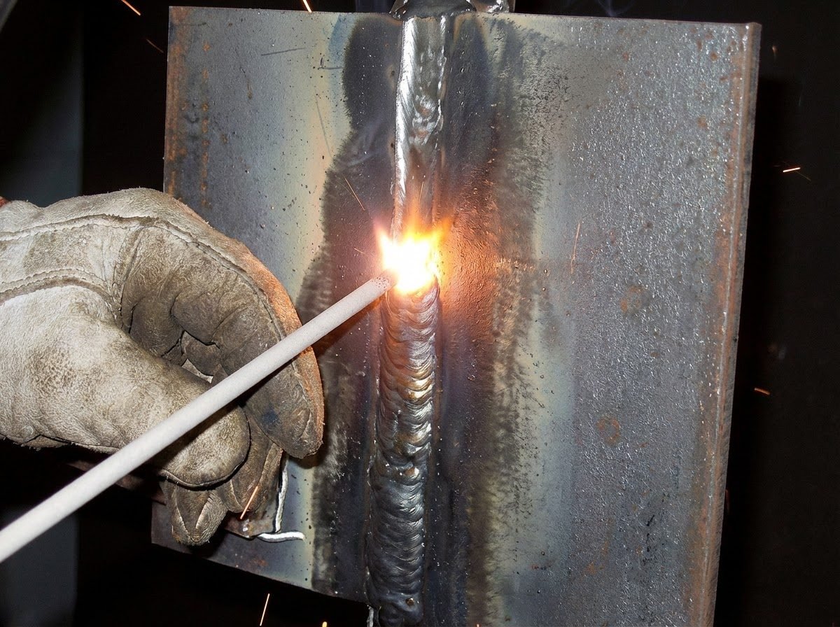 Close-up of leather-gloved hand holding E7018 stick electrode performing structural welding, showing bright arc, consistent weld bead, and heat affected zone on steel plate